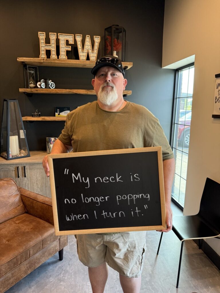 A Health From Within patient holding a chalkboard that reads: My neck is no longer popping when I turn it.