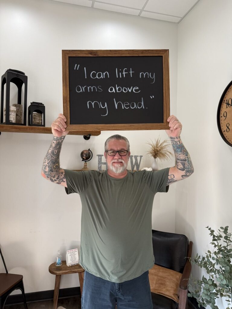 A Health From Within patient holding a chalkboard that reads: I can lift my arms above my head.